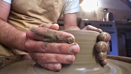 A craftsperson shapes clay on a pottery wheel, hands covered in wet earth. The workshop setting, with a stove in the background, emphasizes the skilled manipulation of clay.