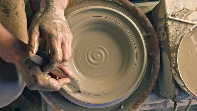 A craftsperson's hands skillfully shape clay on a pottery wheel, creating a spiral pattern. The setting is a workshop, with tools and clay remnants visible.