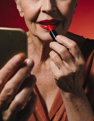 Close-up beauty shot of older woman with wrinkles putting on red lipstick and holding mirror, analog studio shot with film grain structure of female best ager model in her 70s with makeup and red lips