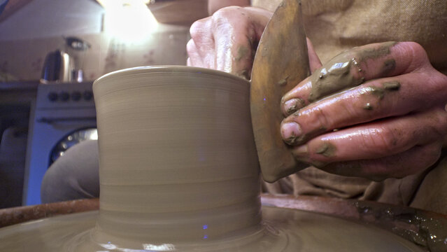 A craftsperson shapes a clay pot on a pottery wheel, their hands skillfully guiding the wet clay. The setting is a workshop, with tools and a stove in the background.