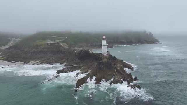Aerial view of the Petit Minou Lighthouse standing tall on a rocky outcrop, waves crashing around it, a beacon against the foggy horizon, Bretagne, France.