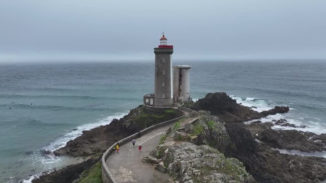 Aerial view of Petit Minou lighthouse amidst rocky outcrops and crashing waves, with people strolling along the causeway, Bretagne, France.