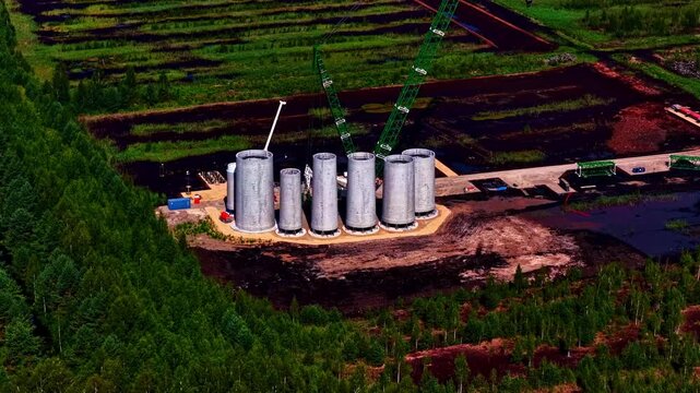 Massive wind-turbine tower sections stand in rows on a remote construction site, surrounded by cranes, machinery and green peat fields as the renewable-energy project rises from the landscape.