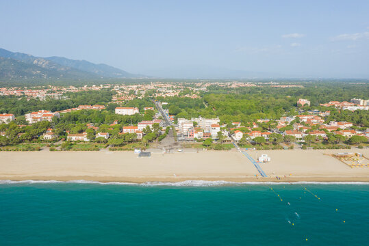 High-angle shot of Argeles with golden sand beach, turquoise water, and a vibrant town bordered by lush greenery and distant mountains under a clear sky. The scene is bright, spacious, and inviting