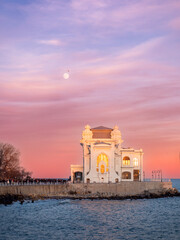 Obraz premium Constanța Casino at sunset, bathed in soft pink and blue tones, with the historic landmark glowing gently above the calm Black Sea, framed by concrete tetrapods in the foreground.