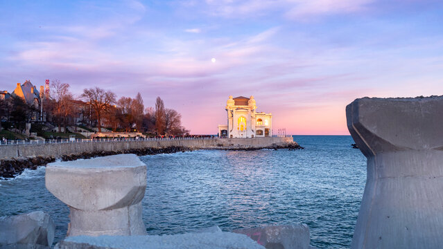 Constanța Casino at sunset, bathed in soft pink and blue tones, with the historic landmark glowing gently above the calm Black Sea, framed by concrete tetrapods in the foreground.