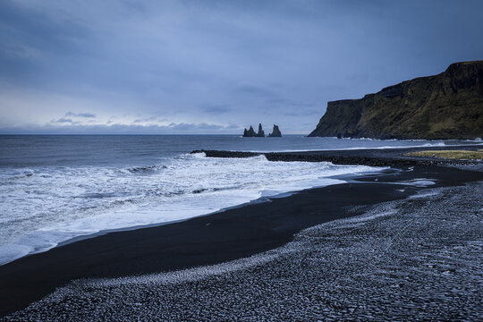 Aerial view of black sand beach meeting the turbulent ocean under a moody sky, with jagged cliffs and the iconic Reynisdrangar sea stacks standing guard, Vik i Myrdal, Iceland.