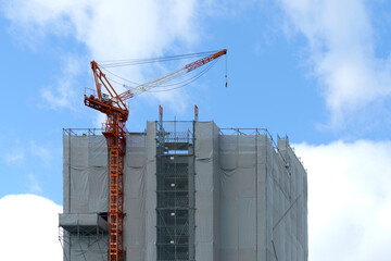 Large orange tower crane on top of a building under construction wrapped in grey safety mesh. Skyscraper development project against a bright blue sky with white clouds. Urban growth concept. © sirawut