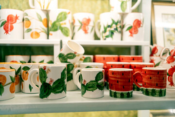Shelf display of handmade ceramic mugs painted with fruit and cactus patterns in a gift shop.