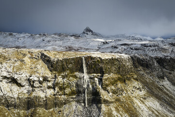 Aerial view of a dramatic waterfall cascading down a steep, snow-dusted cliff face, contrasting with the rugged, snow-covered mountains in the distance, Iceland.