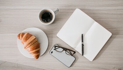 Top view of a light wooden desk with black coffee in a cup a croissant on a plate an open blank notebook with pen eyeglasses and a smartphone arranged neatly for a calm morning workspace