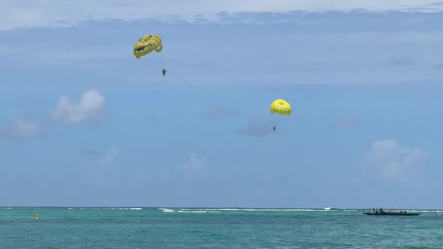 A person is parasailing high in the sky, enjoying the view over a beachfront area. The colorful parachute contrasts with the blue sky and white clouds.