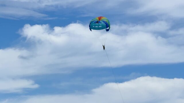 A person is parasailing high in the sky, enjoying the view over a beachfront area. The colorful parachute contrasts with the blue sky and white clouds.
