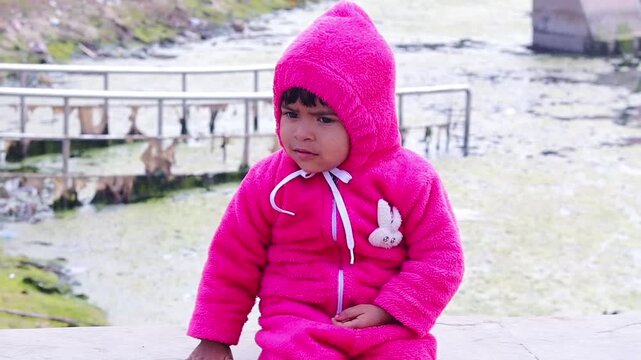 Portrait of two years old Indian baby girl in pink winter wear sitting outdoors near water pond.