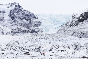 View of a solitary figure in a red coat stands dwarfed by the vast, icy expanse of a glacier nestled between snow-capped mountains, Golden Circle, Iceland.