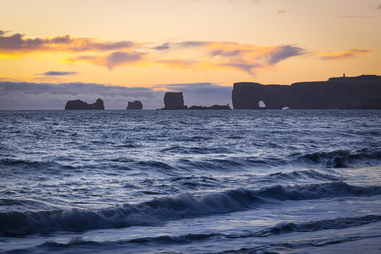 View of the dramatic cliffs meet the churning sea under a fiery sunset, the waves crashing onto the shore in a dance of light and shadow, Golden Circle, Iceland.