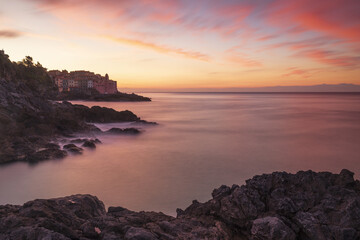 View of the dramatic sunset paints the sky in fiery hues above the tranquil sea, illuminating the rugged coastline and distant buildings, Tellaro, Liguria, Italy.