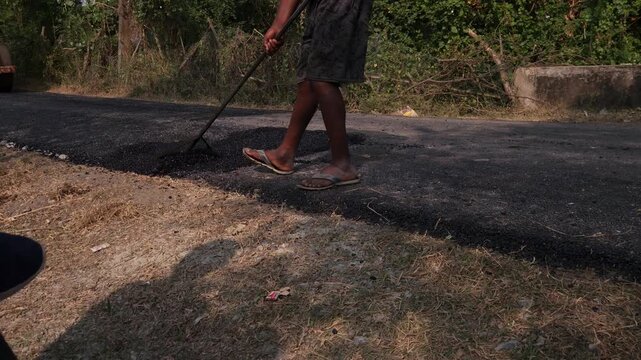 Close-up view of a worker leveling freshly laid bitumen with a rake during road construction, showcasing infrastructure development in a village area.