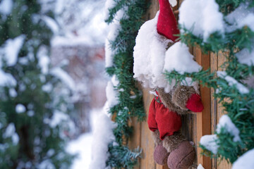 Winter scene featuring a decorative plush toy with a red hat, hanging on a wooden fence adorned with snow-covered greenery, creating a festive holiday atmosphere
