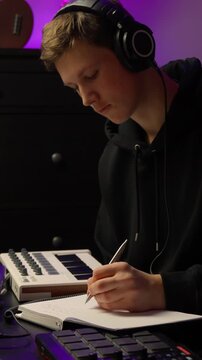 Music producer taking notes in notebook MIDI keyboard and sound pad in dimly lit home studio, vertical