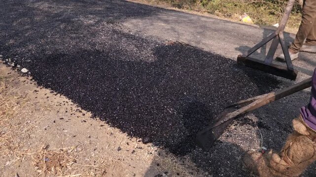 Close-up view of a worker leveling freshly laid bitumen with a rake during road construction, showcasing infrastructure development in a village area.