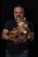 A bearded middle-aged man holding a fluffy ginger cat against a dark studio background..
