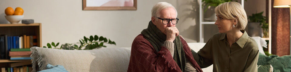 Header of Senior Caucasian man sitting on sofa coughing into fist, while middle aged Caucasian...