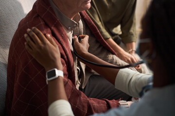Fototapeta premium Middle aged Black woman using stethoscope examining chest of senior Caucasian man sitting on couch while another observing, hands and partial faces visible, focus on medical care interaction