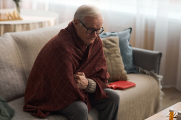 Senior Caucasian man sitting on sofa wrapped in blanket, holding arms close to chest, appearing...