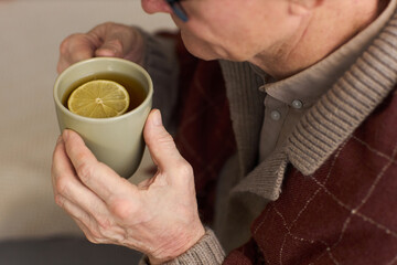 Senior Caucasian man holding mug with hot beverage and lemon slice, sitting indoors, wearing glasses and sweater, hands visible, looking down at drink, close up view