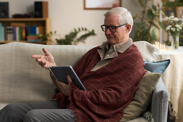 Senior Caucasian man sitting on sofa using digital tablet, gesturing with hand, wearing glasses and blanket, engaged in video call in home living room setting