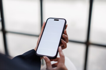 View of a woman sits indoors and holds a smartphone with a white screen mockup, with blurred background. people and wireless technology concepts
