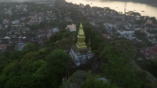 Aerial drone footage of Mount Phousi (Phou Si) rising above the historic city of Luang Prabang, Laos. The sacred hill is crowned by a golden Buddhist stupa and surrounded by temples, traditional rooft
