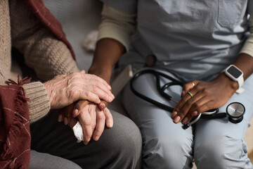 Fototapeta premium Senior Caucasian man sitting with hands resting on lap, being comforted by young adult Black woman holding stethoscope, showing support and care in medical setting