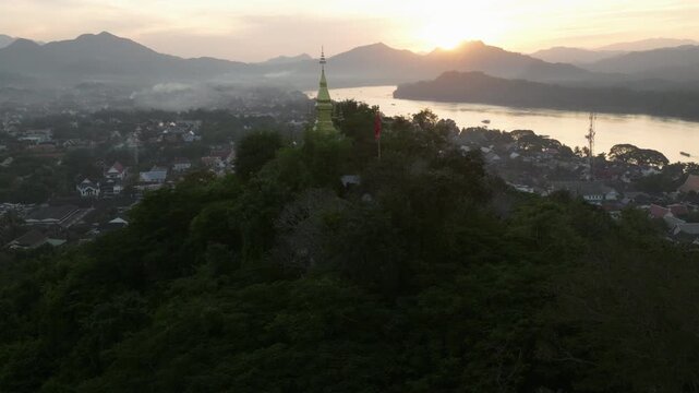 Aerial drone footage of Mount Phousi (Phou Si) rising above the historic city of Luang Prabang, Laos. The sacred hill is crowned by a golden Buddhist stupa and surrounded by temples, traditional rooft