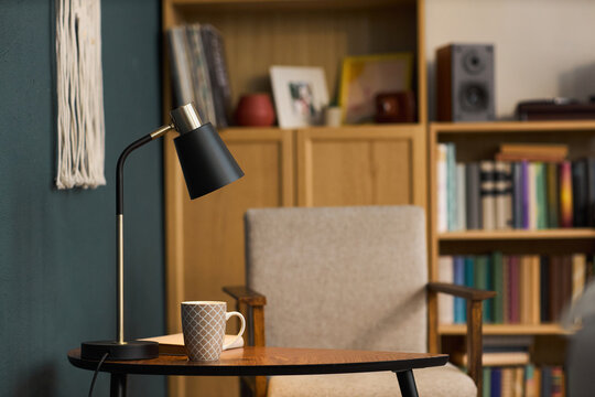 Modern table lamp standing on wooden side table next to patterned coffee mug in cozy reading corner, with bookshelf filled with books and framed photos in background