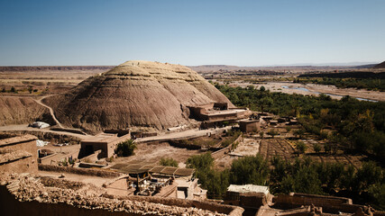 Wide view of Ait Ben Haddou area in Morocco, with a rounded desert hill, earthen buildings, palm trees, fields, and a dry river valley under clear blue sky.