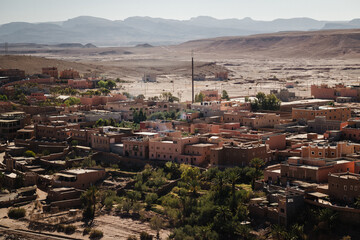 Wide view of Ait Ben Haddou in Morocco, showing earthen houses with flat roofs, palm trees, and a dry valley, with desert hills and distant mountains under clear daylight.