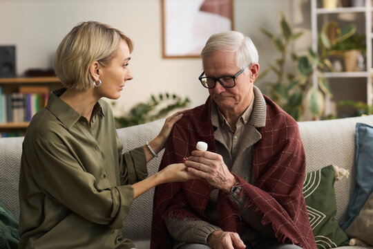 Caucasian middle aged woman supporting senior Caucasian man by handing medication bottle while sitting together on sofa, woman gently touching man's shoulder, plants in background
