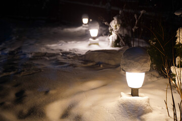 Snow-covered pathway illuminated by outdoor lamps at night, creating a serene winter atmosphere with soft light reflecting on the white snow, enhancing the tranquil scene