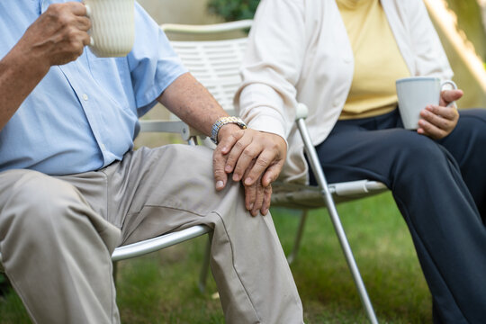 Close-up of happy Asian elderly couple holding hands while sitting on chair and drinking coffee or tea in garden home. Old senior marriage care together. - Powered by Adobe