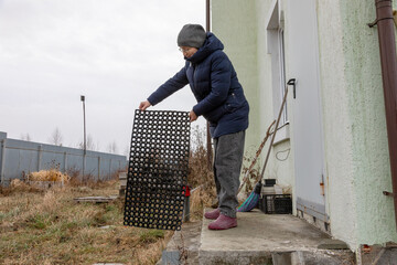 Woman shaking dirt from a rubber mat on the porch of a country house, performing outdoor cleaning chores during autumn or spring season.