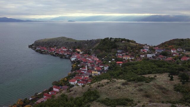 Aerial video of Lin village on Lake Ohrid in Albania, with shoreline houses and soft sun-rays breaking through the clouds.