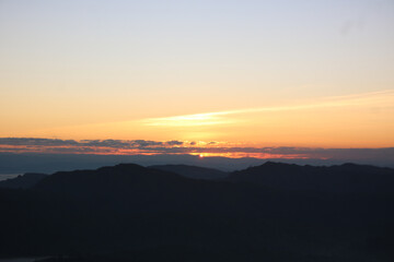 Fototapeta premium Dawn's Embrace over Mountains: The golden sun ascends, casting its warm glow upon the silhouettes of majestic mountains. The tranquil scene evokes a sense of peace and the promise of a new day.