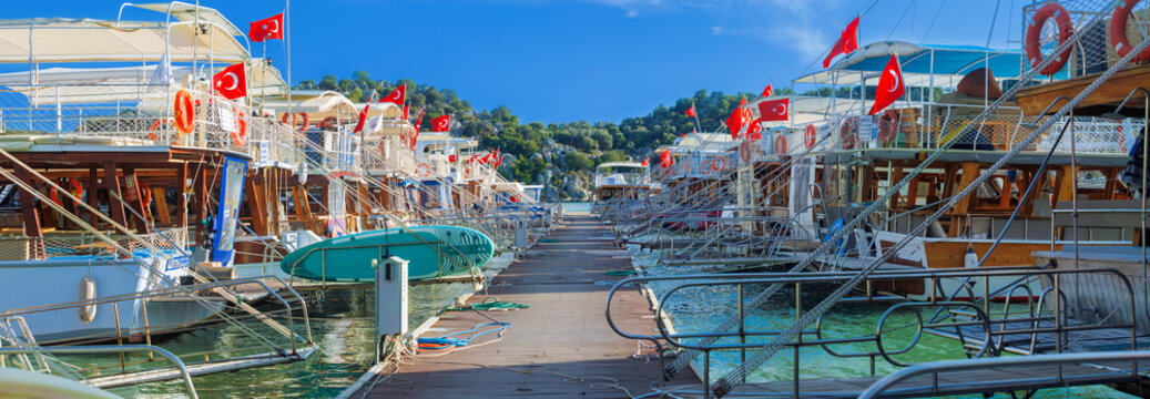 Panoramic wooden pier walkway lined with traditional Turkish gulets and boats flying red flags in a turquoise marina bay. Serene summer scene with green hills and blue skies,