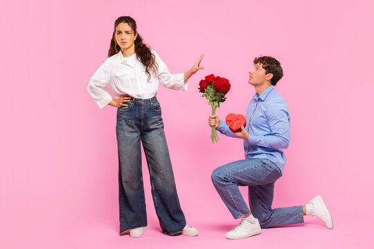 Guy standing on one knee with flowers and gift for Valentine's Day while offended lady rejecting him, standing on pink background