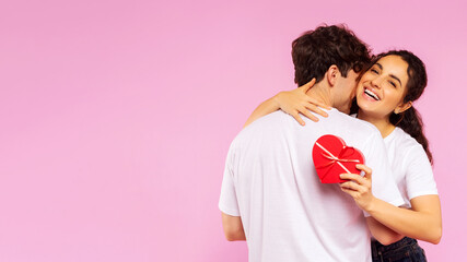 Happy couple in love celebrating Valentine's day, lady holding heart shaped gift box while embracing boyfriend, pink studio background