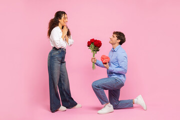 Romantic European guy giving his beloved girlfriend flowers and gift for Valentine's Day over pink studio background, full length shot