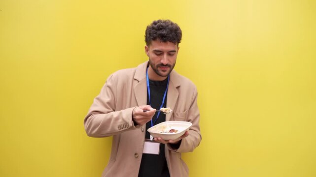 Young man wearing a blazer and an id badge having a lunch break against a vibrant yellow wall. He is enjoying his takeaway meal