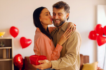 Woman kissing man, giving gift box, couple embracing in room filled with red heart-shaped balloons, celebrating Valentines day or anniversary